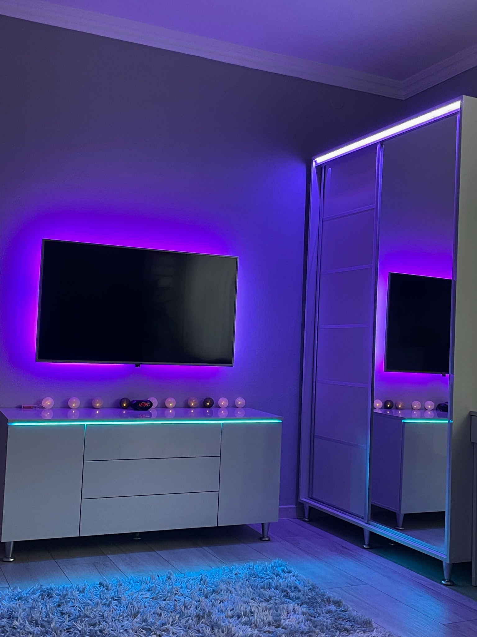 Pokój z szafą przesuwną z lustrami, komodą i telewizorem podświetlonymi ledowym światłem w odcieniach fioletu i turkusu, dywan shaggy na podłodze.