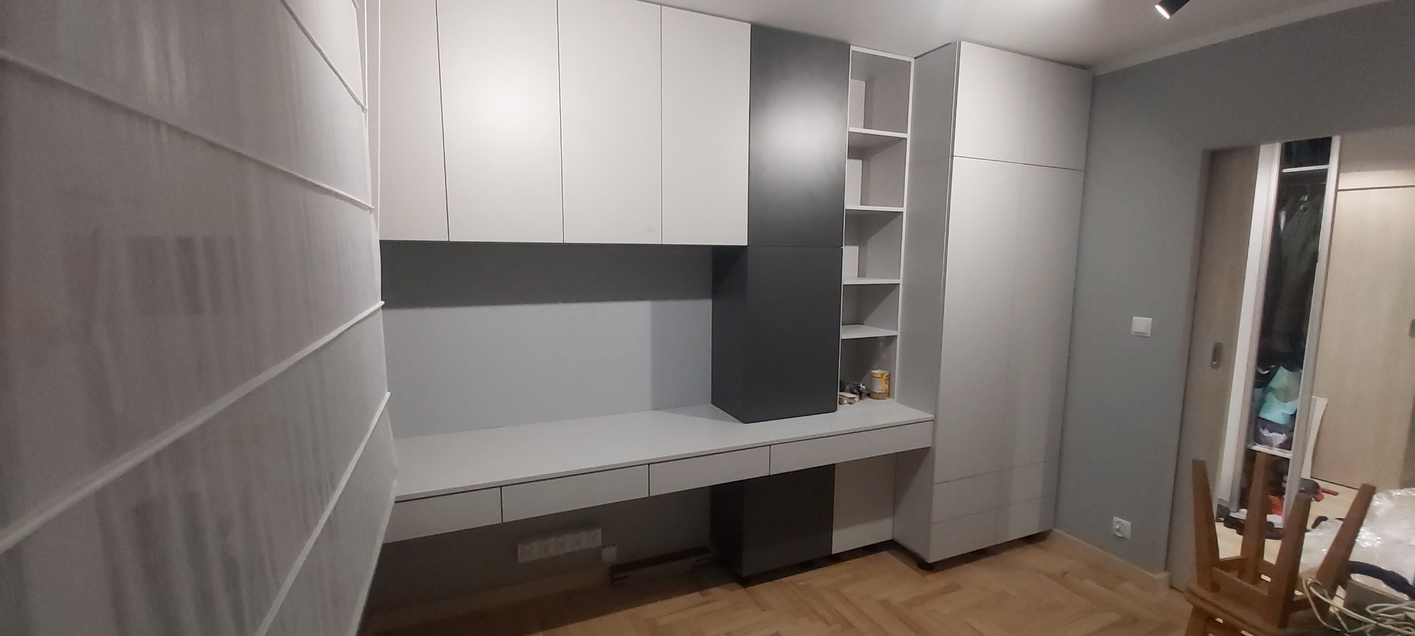 Nowoczesny pokój z zabudową meblową: białe szafki wiszące, szara szafa z półkami i biurko z szufladami, uzupełnione roletą rzymską na oknie.