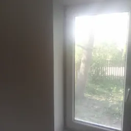 Świeżo pomalowana ściana w odcieniu szarości, sąsiadująca z oknem PCV, przez kt&oacute;re widać drzewo i ogrodzenie. Na parapecie leżą ż&oacute;łte ściereczki i szary bloczek szlifierski.