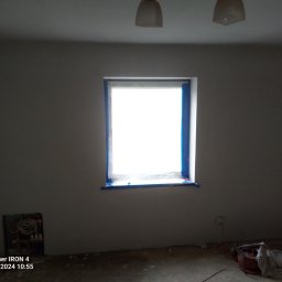 Sławomir kak - Pokój w trakcie przygotowań do malowania: okno zabezpieczone niebieską taśmą malarską, ściany po wstępnym wyrównaniu, widoczne instalacje grzewcze oraz zwinięty przedłużacz na podłodze.