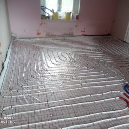 Sławomir kak - Rozłożone rury ogrzewania podłogowego na folii z siatką w pomieszczeniu w trakcie remontu, widoczne niedomalowane ściany i okno z akcesoriami malarskimi.