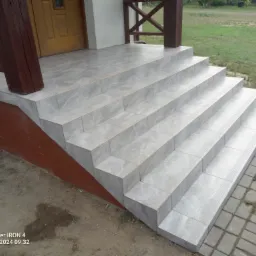 Zewnętrzne schody wejściowe do domu wyłożone szarymi płytkami imitującymi marmur, z brązowymi drewnianymi słupkami i balustradą, obok kostka brukowa.