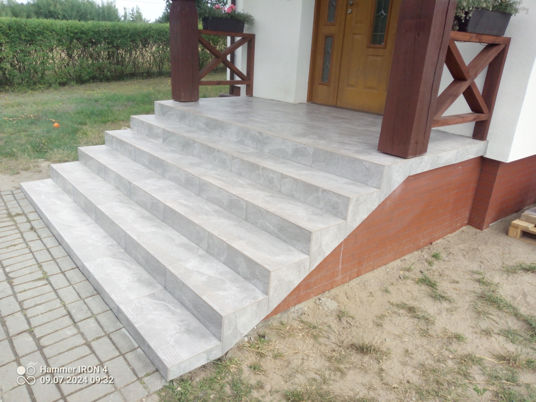 Wyłożone szarymi płytkami schody wejściowe do domu z drewnianą balustradą i kwiatami w donicach.