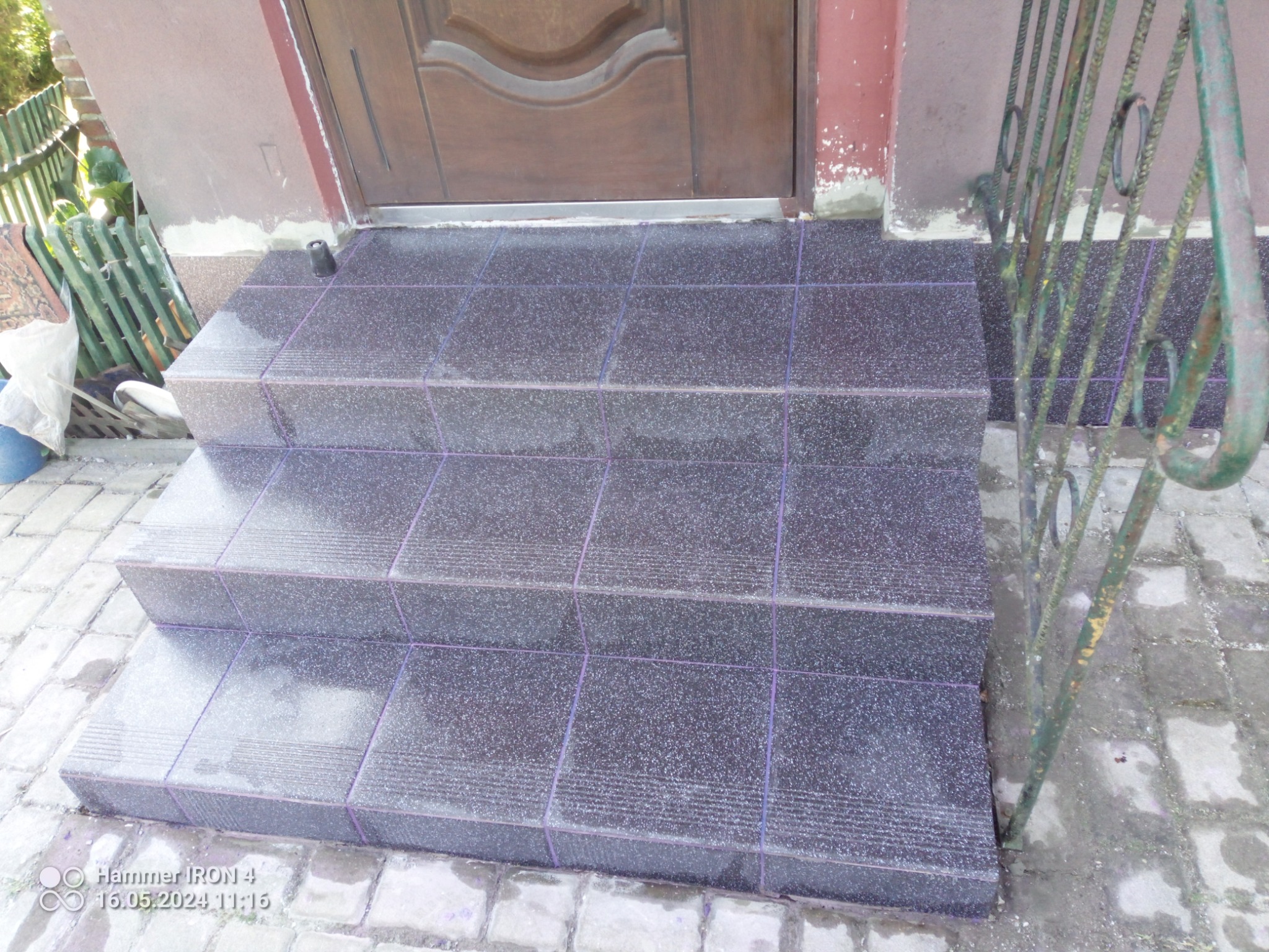 Trzy stopnie wejściowe wyłożone ciemnymi płytkami z jasnym wzorem, widoczne fioletowe fugi, obok zardzewiała barierka, podłoże wyłożone kostką brukową.