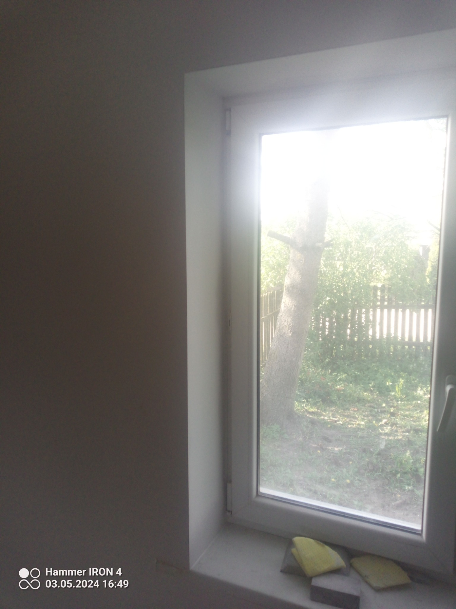 Świeżo pomalowana ściana w odcieniu szarości, sąsiadująca z oknem PCV, przez które widać drzewo i ogrodzenie. Na parapecie leżą żółte ściereczki i szary bloczek szlifierski.