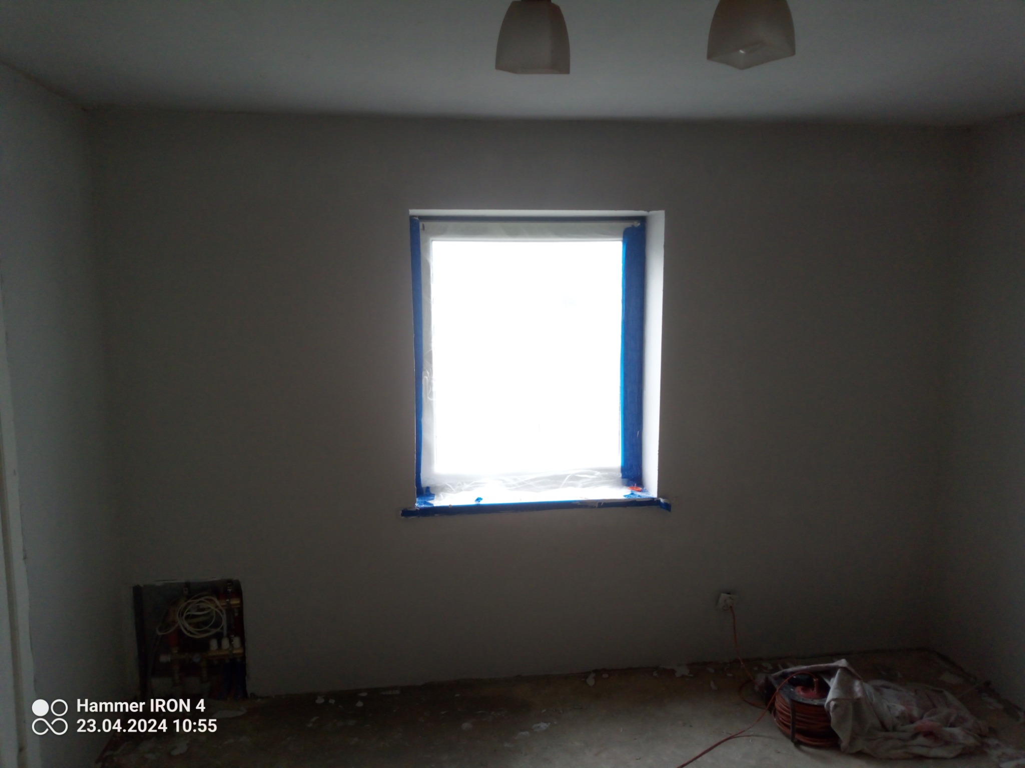 Pokój w trakcie przygotowań do malowania: okno zabezpieczone niebieską taśmą malarską, ściany po wstępnym wyrównaniu, widoczne instalacje grzewcze oraz zwinięty przedłużacz na podłodze.