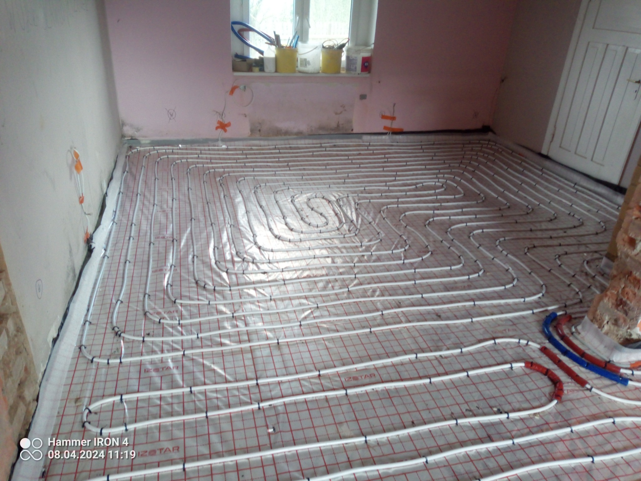 Rozłożone rury ogrzewania podłogowego na folii z siatką w pomieszczeniu w trakcie remontu, widoczne niedomalowane ściany i okno z akcesoriami malarskimi.
