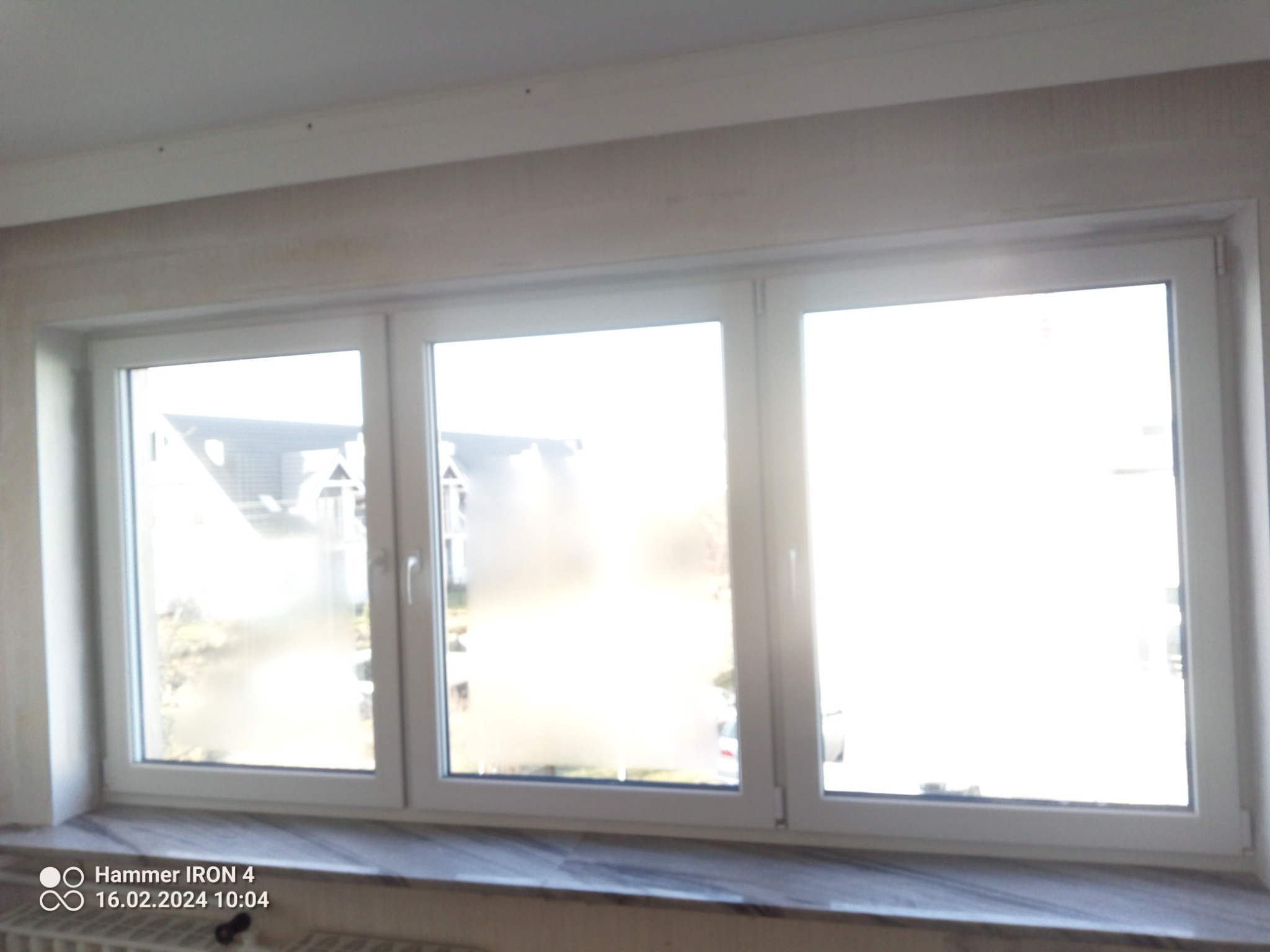 Trzyczęściowe białe okno PCV z widokiem na zewnątrz, zamontowane w jasnym pomieszczeniu, z widocznym fragmentem parapetu i grzejnika.