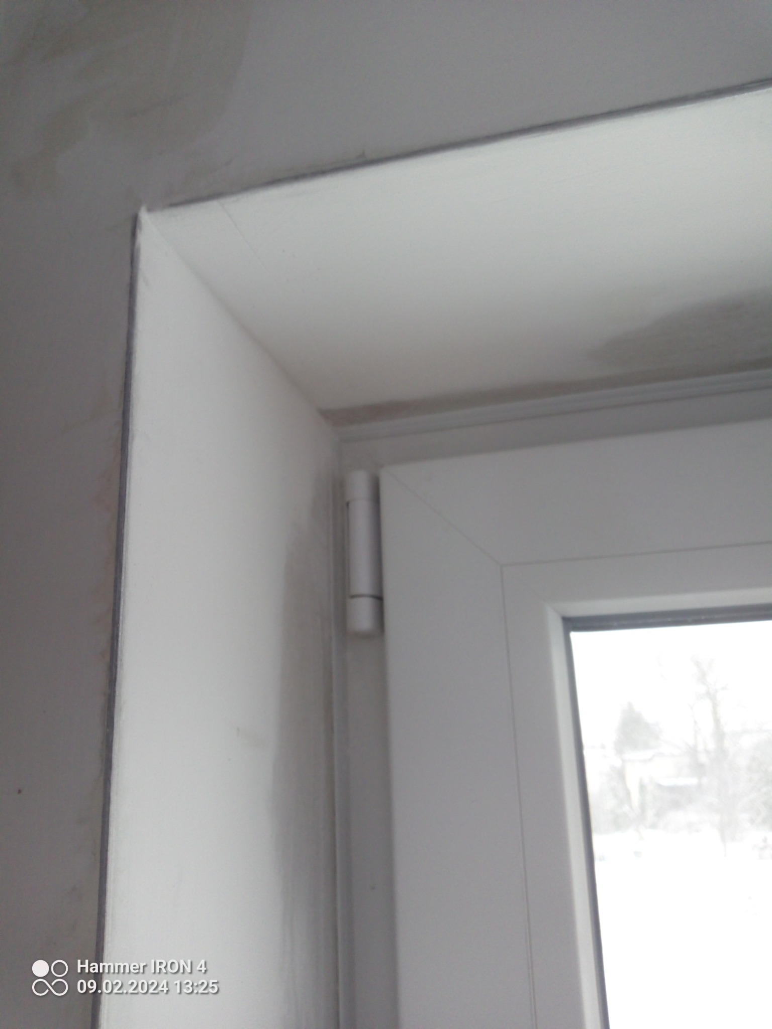 Fragment ściany i okna z widocznymi śladami prac wykończeniowych, na ścianie widoczne niedoskonałości i pozostałości po gładzi, okno białe plastikowe z widocznym zawiasem.