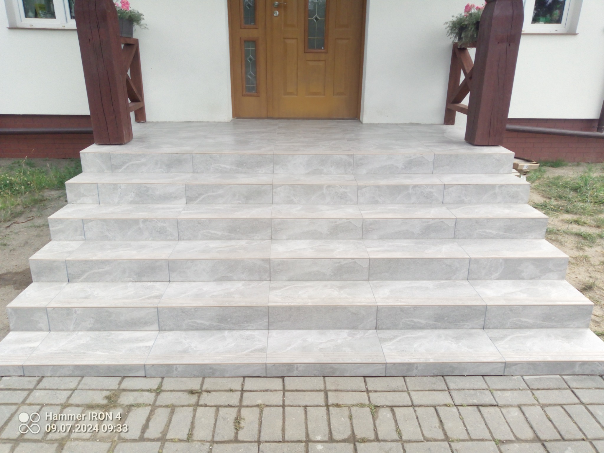 Jasnoszare płytki imitujące marmur na schodach wejściowych do domu, z drewnianą balustradą i kwiatami w doniczkach, widok z dołu na drzwi wejściowe.