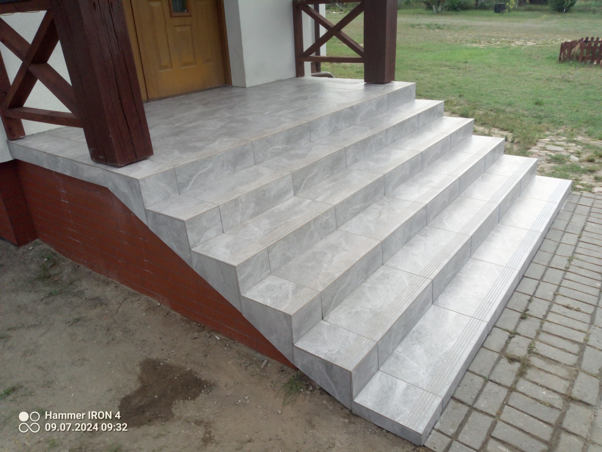 Zewnętrzne schody wejściowe do domu wyłożone szarymi płytkami imitującymi marmur, z brązowymi drewnianymi słupkami i balustradą, obok kostka brukowa.