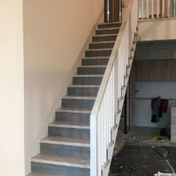 Schody drewniane z szarymi stopniami i białą balustradą, widoczne zabezpieczenia podłogi podczas montażu, fragment kuchni w tle.