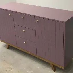 Fioletowa komoda z trzema szufladami i dwojgiem drzwi z geometrycznym wzorem jodełki, umieszczona na drewnianej podstawie z ukośnymi nogami i złotymi gałkami, na tle surowej podłogi w trakcie remontu.