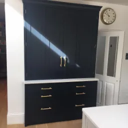 Ciemnogranatowa zabudowa kuchenna z szafkami i szufladami, złote uchwyty, biały blat, drewniana podłoga i wpadające światło słoneczne.