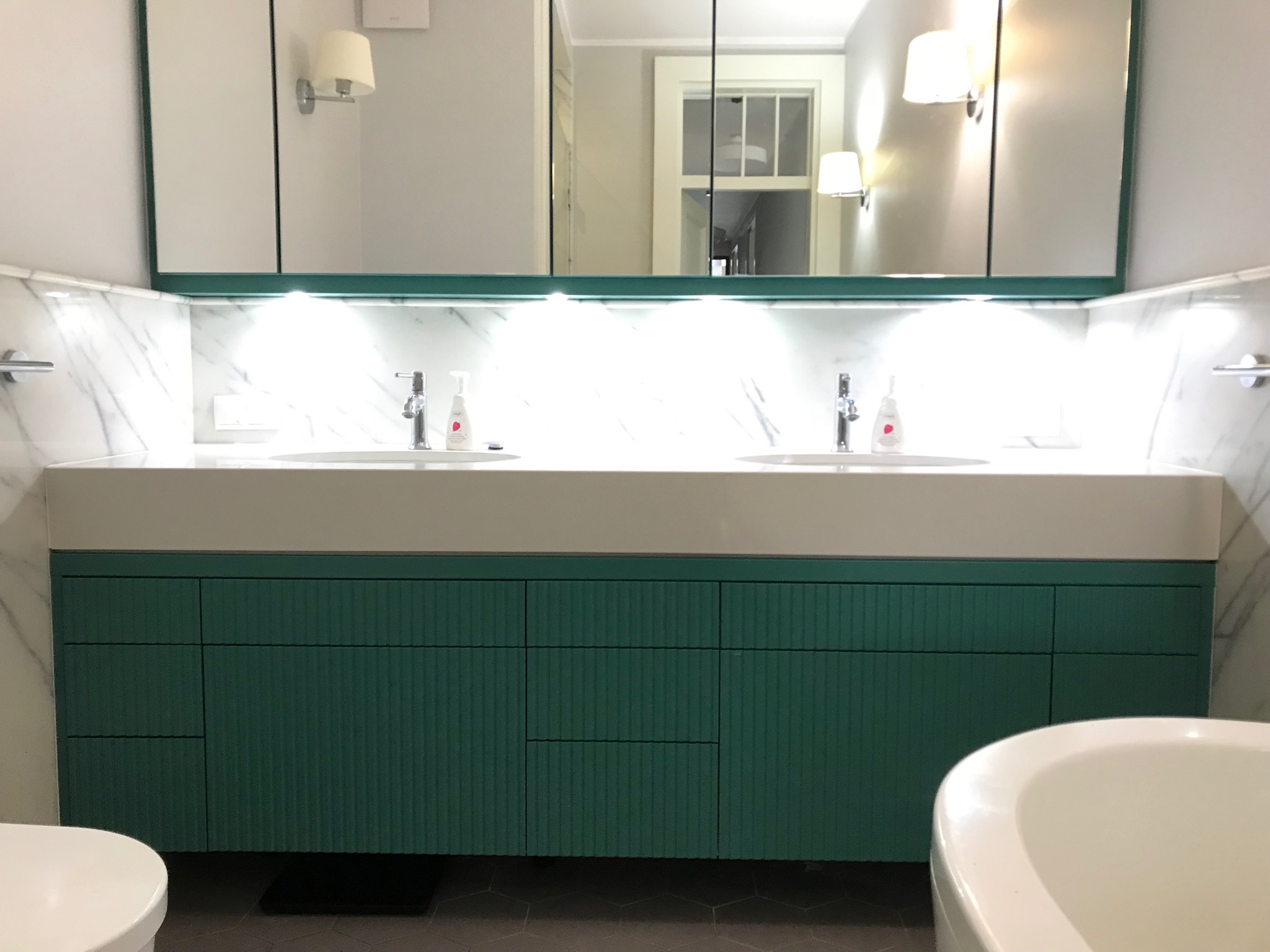 Nowoczesna łazienka z podwójną umywalką na szafce w kolorze butelkowej zieleni, marmurowym blatem i podświetleniem LED, widok z perspektywy wanny.