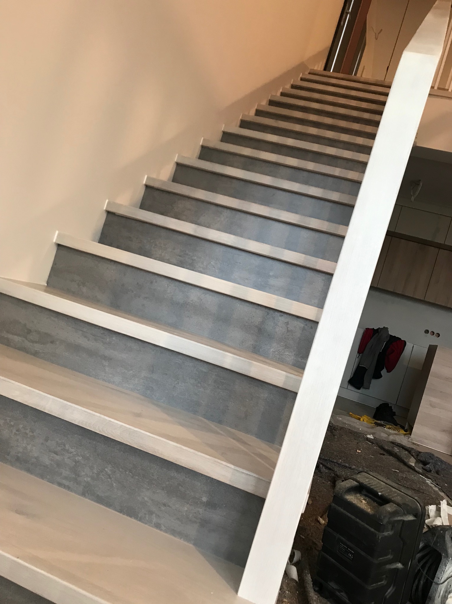 Schody wewnętrzne z szarymi stopniami i białą balustradą, widok od dołu, w tle widoczny fragment wnętrza z szafkami i ubraniami, na podłodze widoczne narzędzia i akcesoria budowlane.