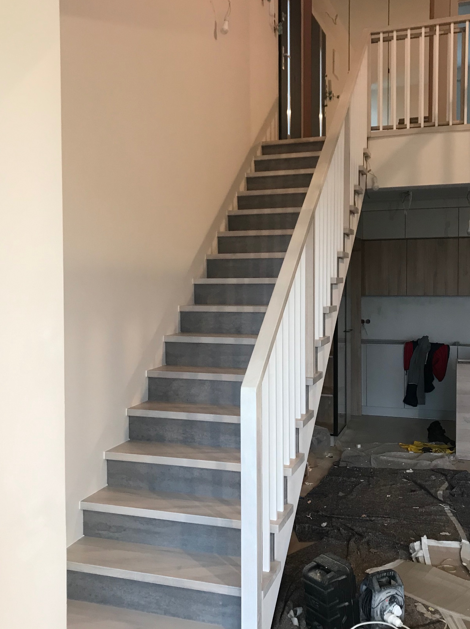 Schody drewniane z szarymi stopniami i białą balustradą, widoczne zabezpieczenia podłogi podczas montażu, fragment kuchni w tle.