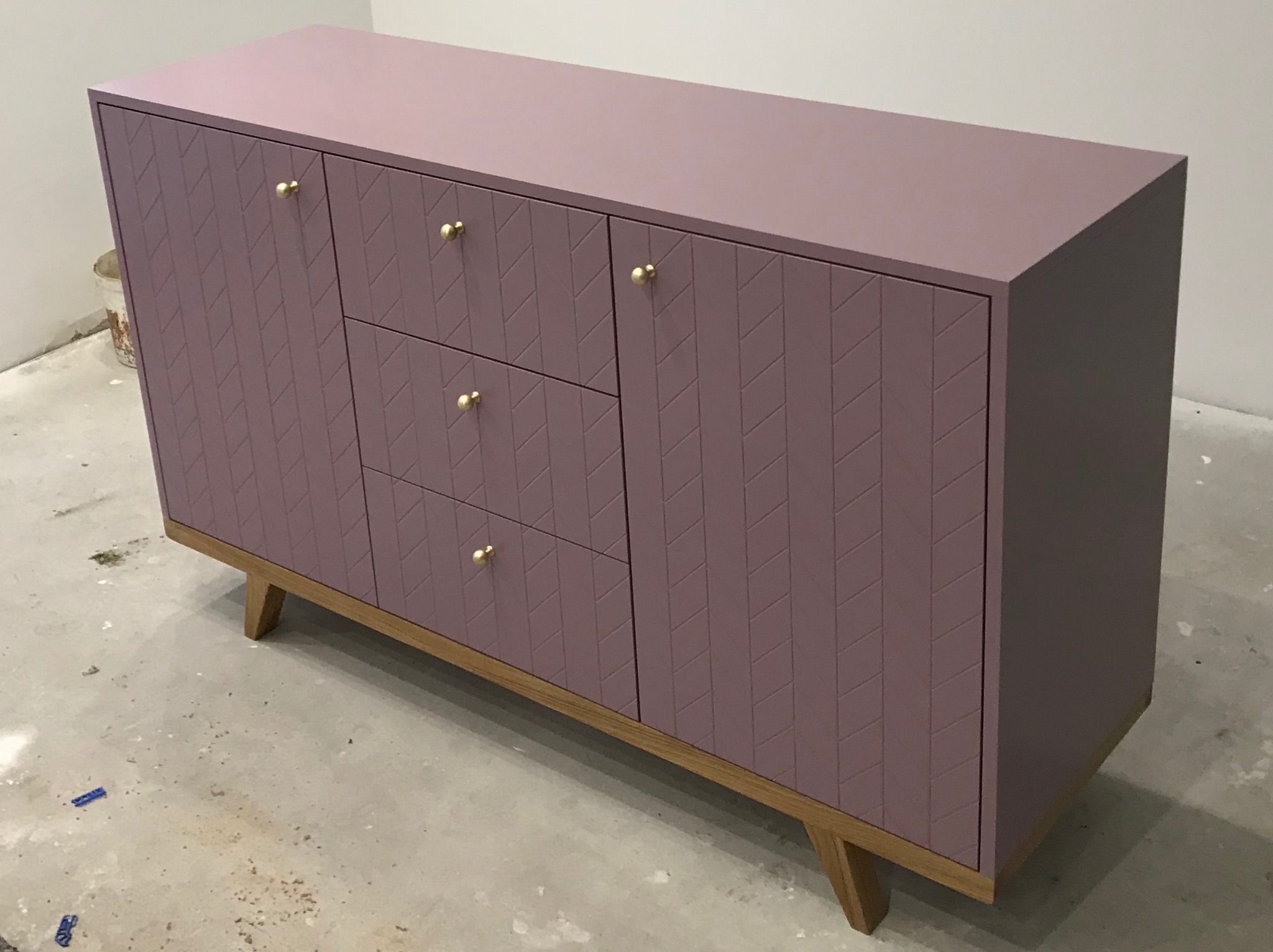 Fioletowa komoda z trzema szufladami i dwojgiem drzwi z geometrycznym wzorem jodełki, umieszczona na drewnianej podstawie z ukośnymi nogami i złotymi gałkami, na tle surowej podłogi w trakcie remontu.