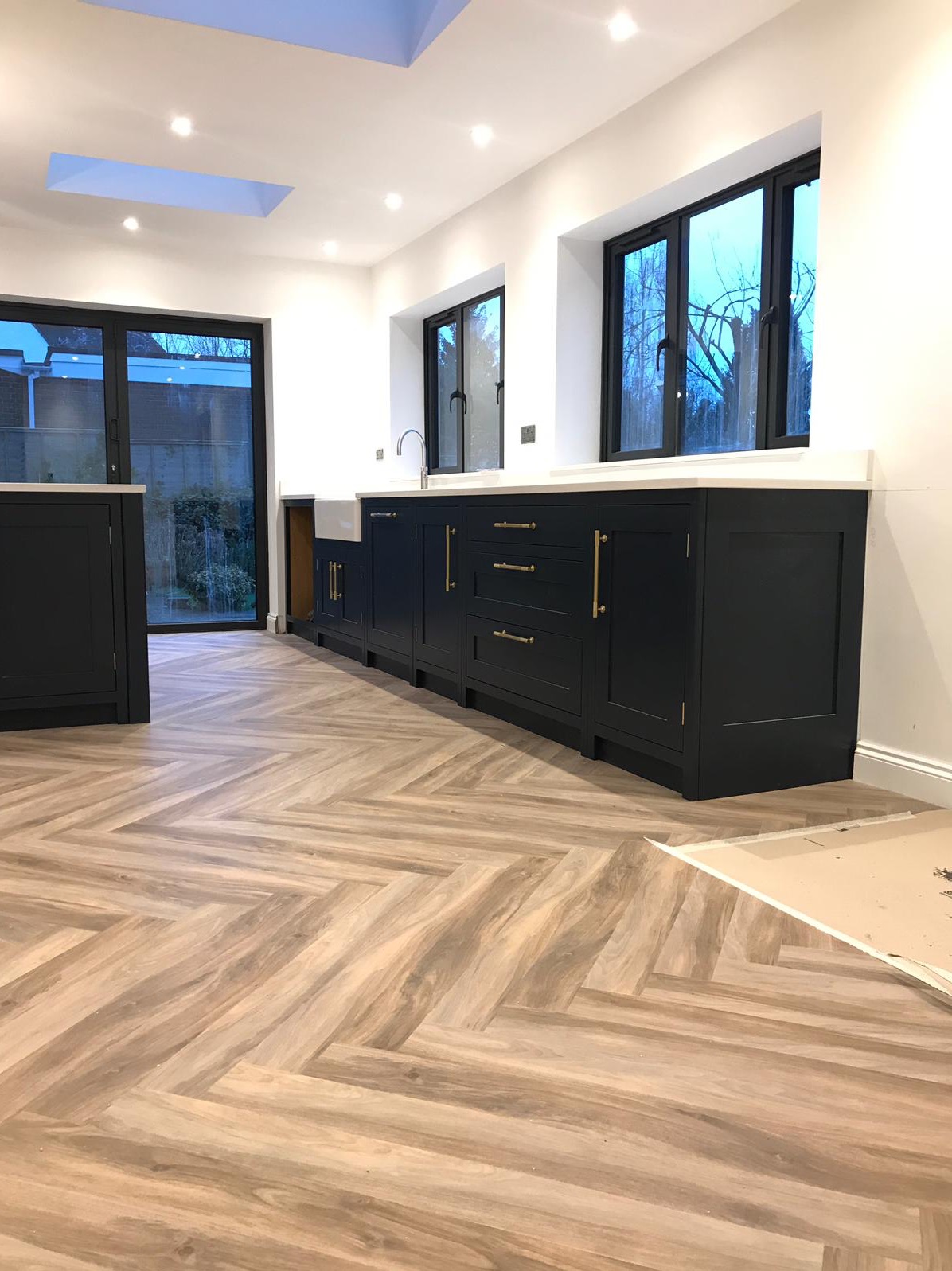 Nowoczesna kuchnia z ciemnymi szafkami, złotymi uchwytami i jasnym blatem, widok na zewnątrz przez okno i drzwi balkonowe, podłoga ułożona w jodełkę.