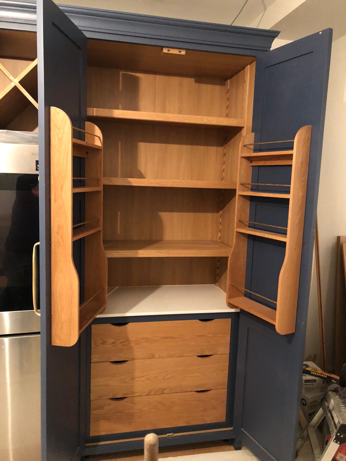 Otwarta, dwudrzwiowa szafa w kolorze granatowym z drewnianymi półkami i szufladami w dolnej części; widoczne listwy na drzwiach, biały blat w środku, a w tle lodówka ze stali nierdzewnej.