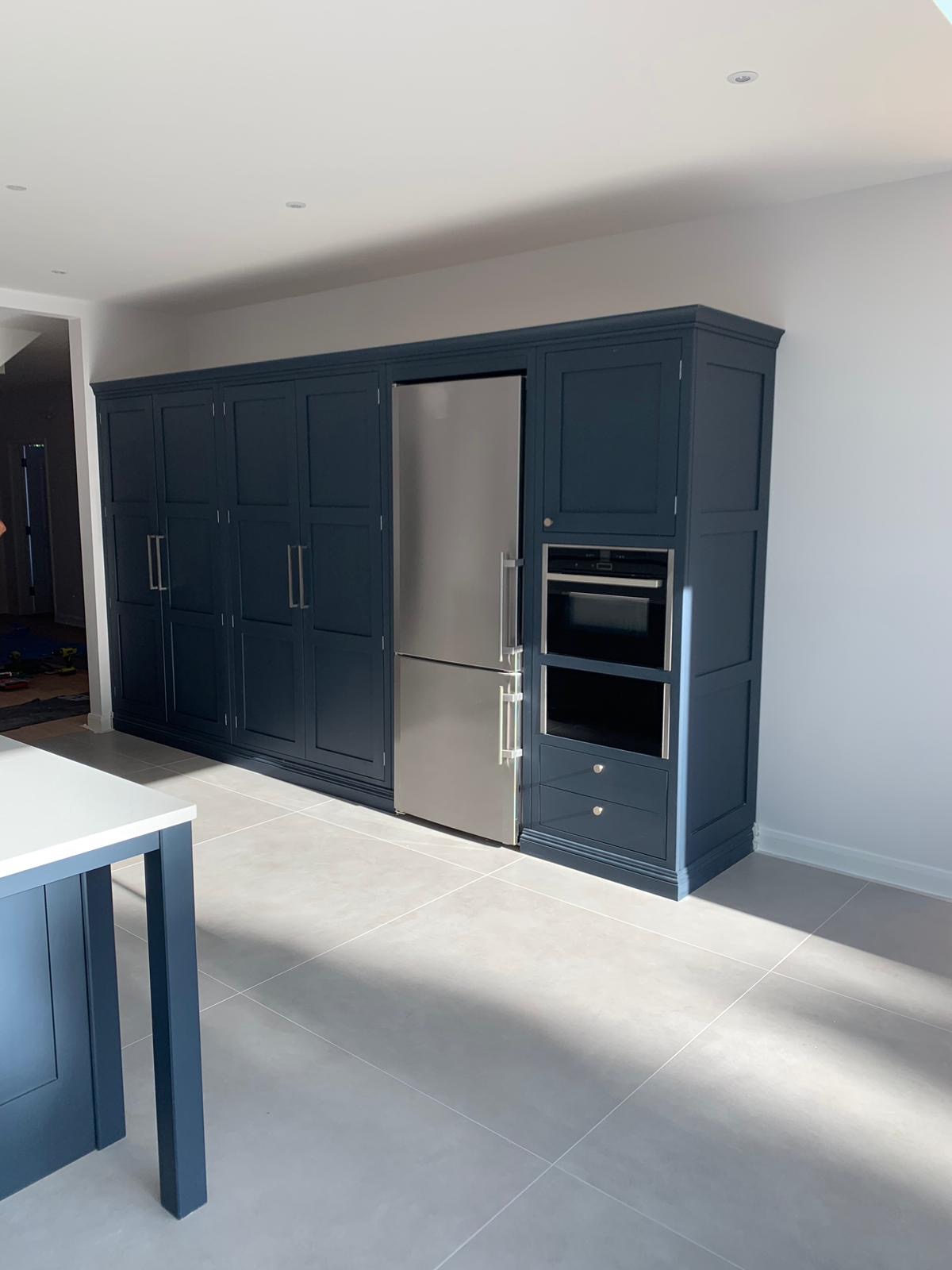 Granatowa zabudowa kuchenna z lodówką i piekarnikiem, w tle widoczne szafki, całość w nowoczesnym wnętrzu z szarymi płytkami podłogowymi i fragmentem wyspy kuchennej.