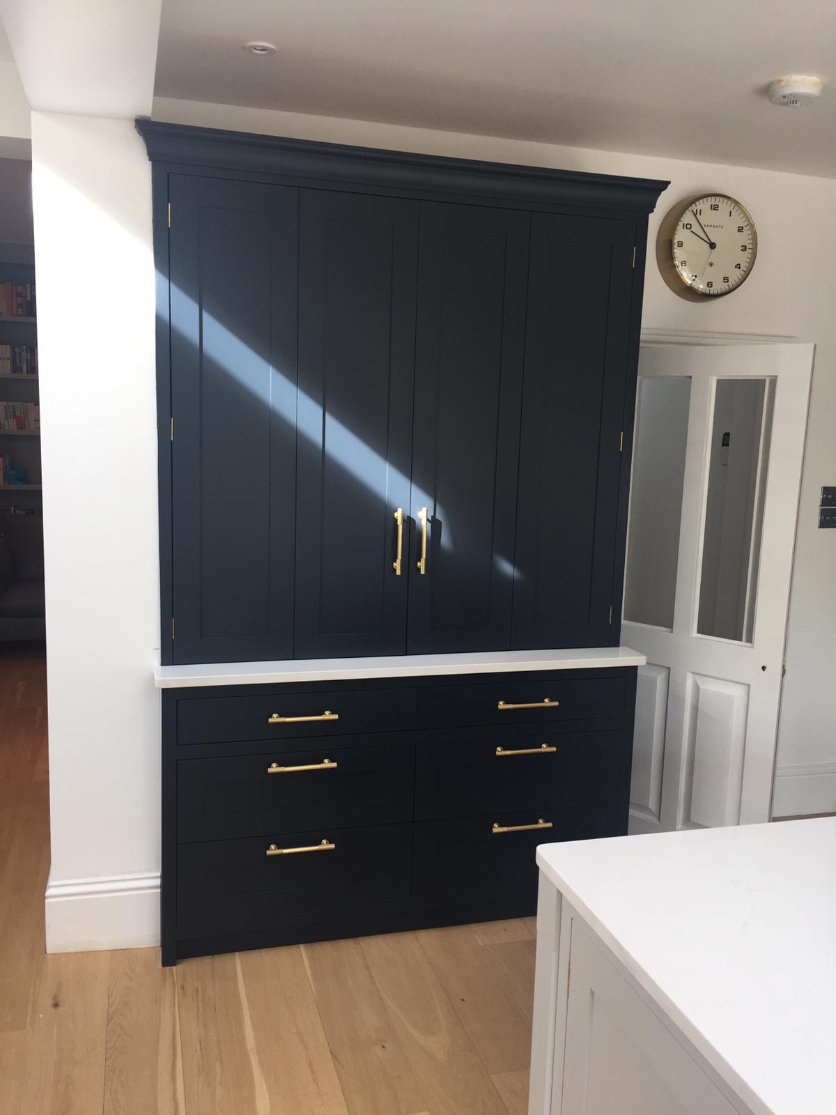 Ciemnogranatowa zabudowa kuchenna z szafkami i szufladami, złote uchwyty, biały blat, drewniana podłoga i wpadające światło słoneczne.