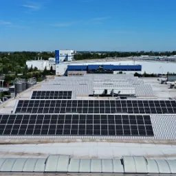 Widok z góry na rozległy dach przemysłowy z panelami słonecznymi, z budynkami fabrycznymi i zielenią w tle pod błękitnym niebem.