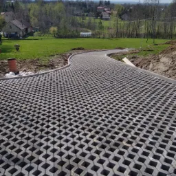 Ułożona, ażurowa nawierzchnia z betonowych płyt na podjeździe, widok z góry, w tle zielony krajobraz z domami.