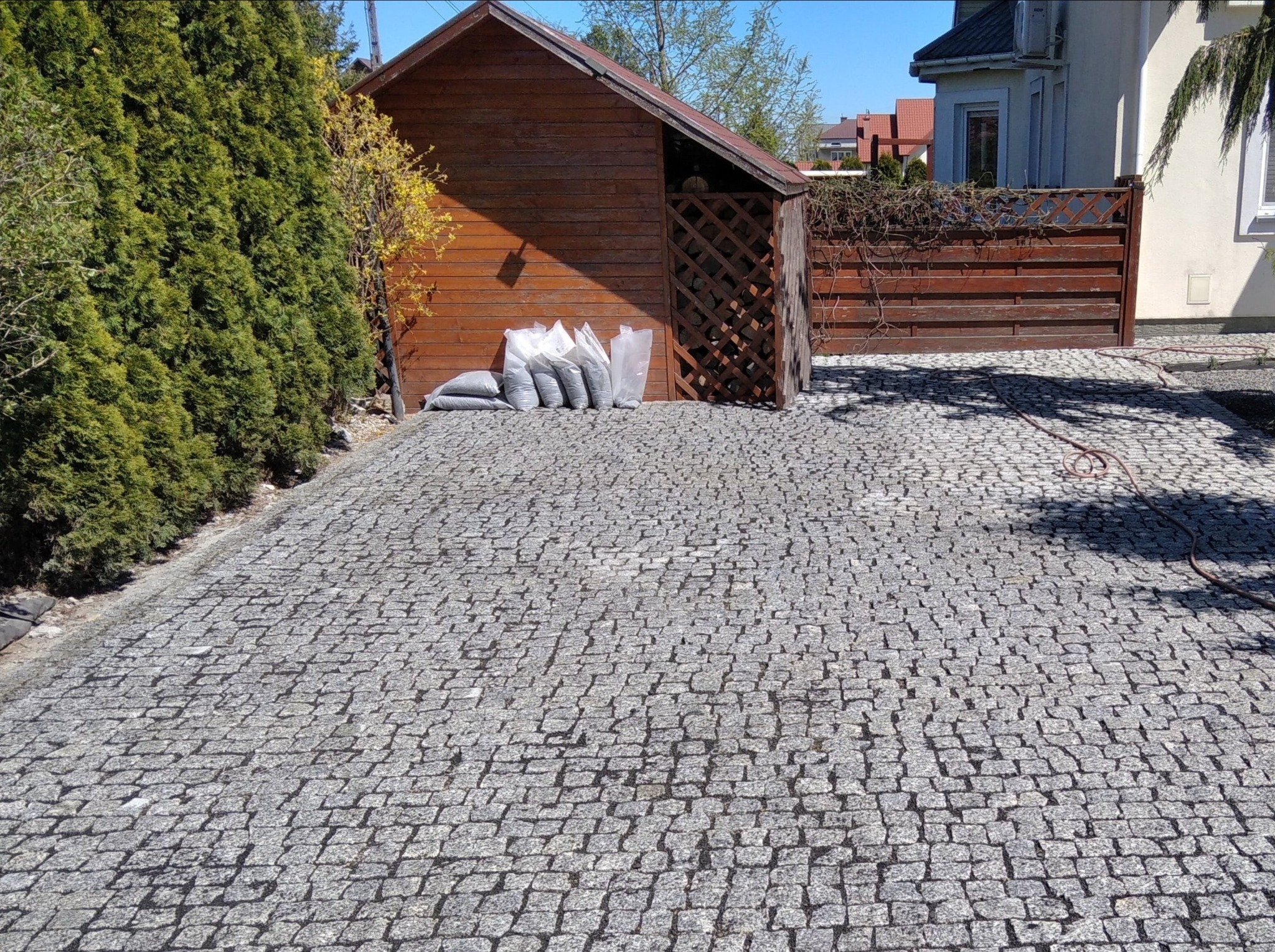 Ułożona szara kostka na podjeździe, przy drewnianej szopie i tujach, obok leżą worki z materiałem.