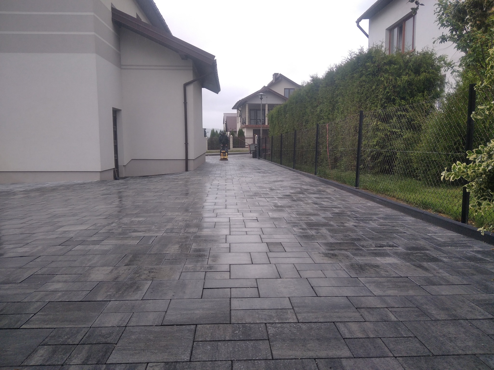 Świeżo ułożona, szara kostka brukowa prowadząca do domu, z widocznym pracownikiem zagęszczającym powierzchnię w oddali.