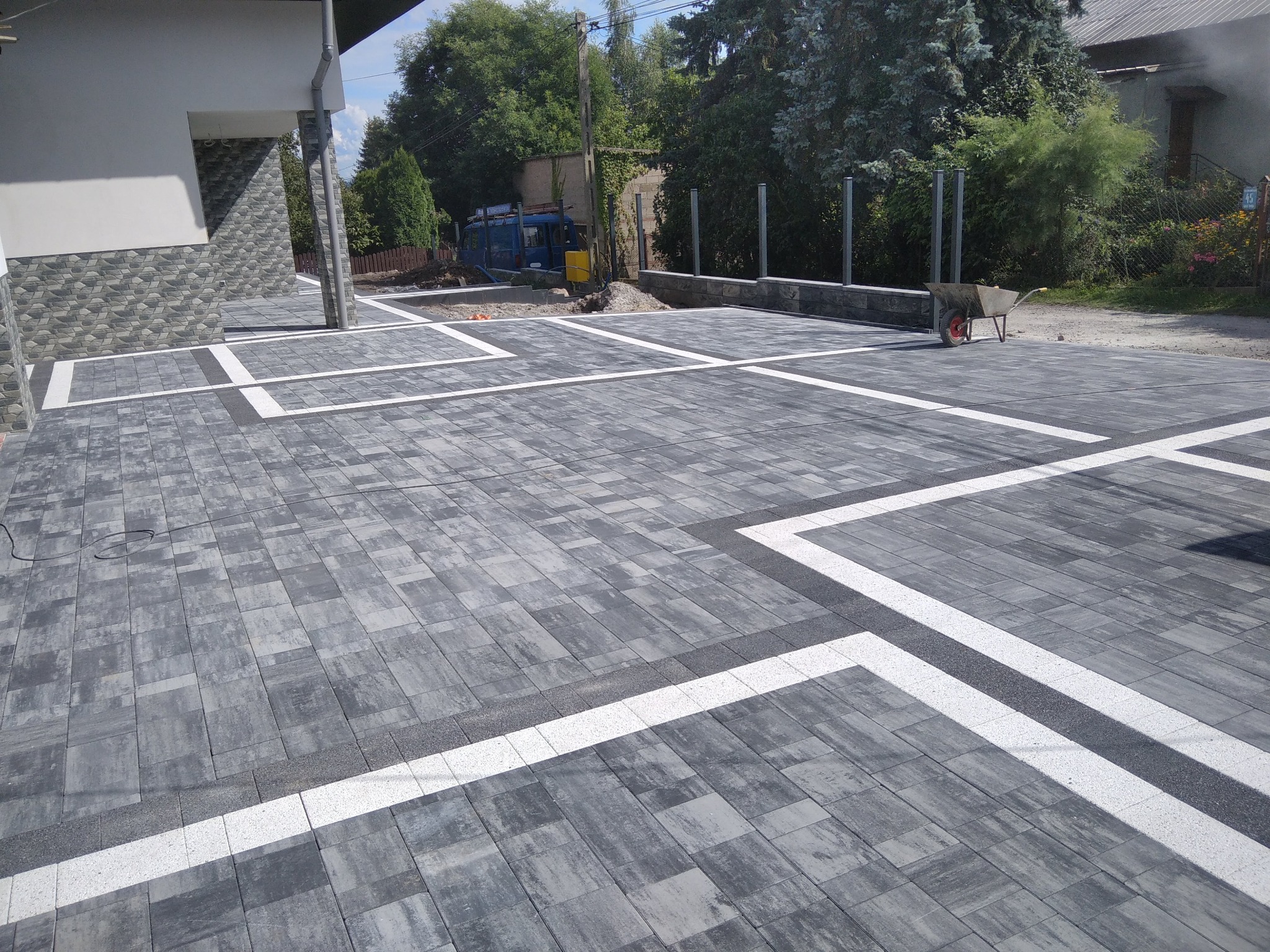 Ułożona szara kostka brukowa z białymi i ciemnoszarymi wstawkami tworząca geometryczny wzór na podjeździe, w tle budynek i częściowo przygotowane podłoże pod dalszą część nawierzchni.