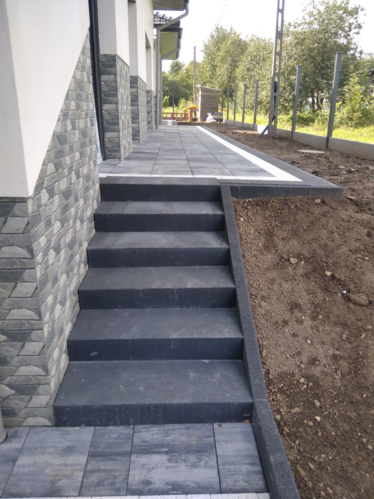 Nowe, ciemnoszare schody z kostki brukowej prowadzące do domu z elewacją wykończoną kamieniem dekoracyjnym, widok na świeżo ułożony chodnik i obrzeża trawnika w tle.