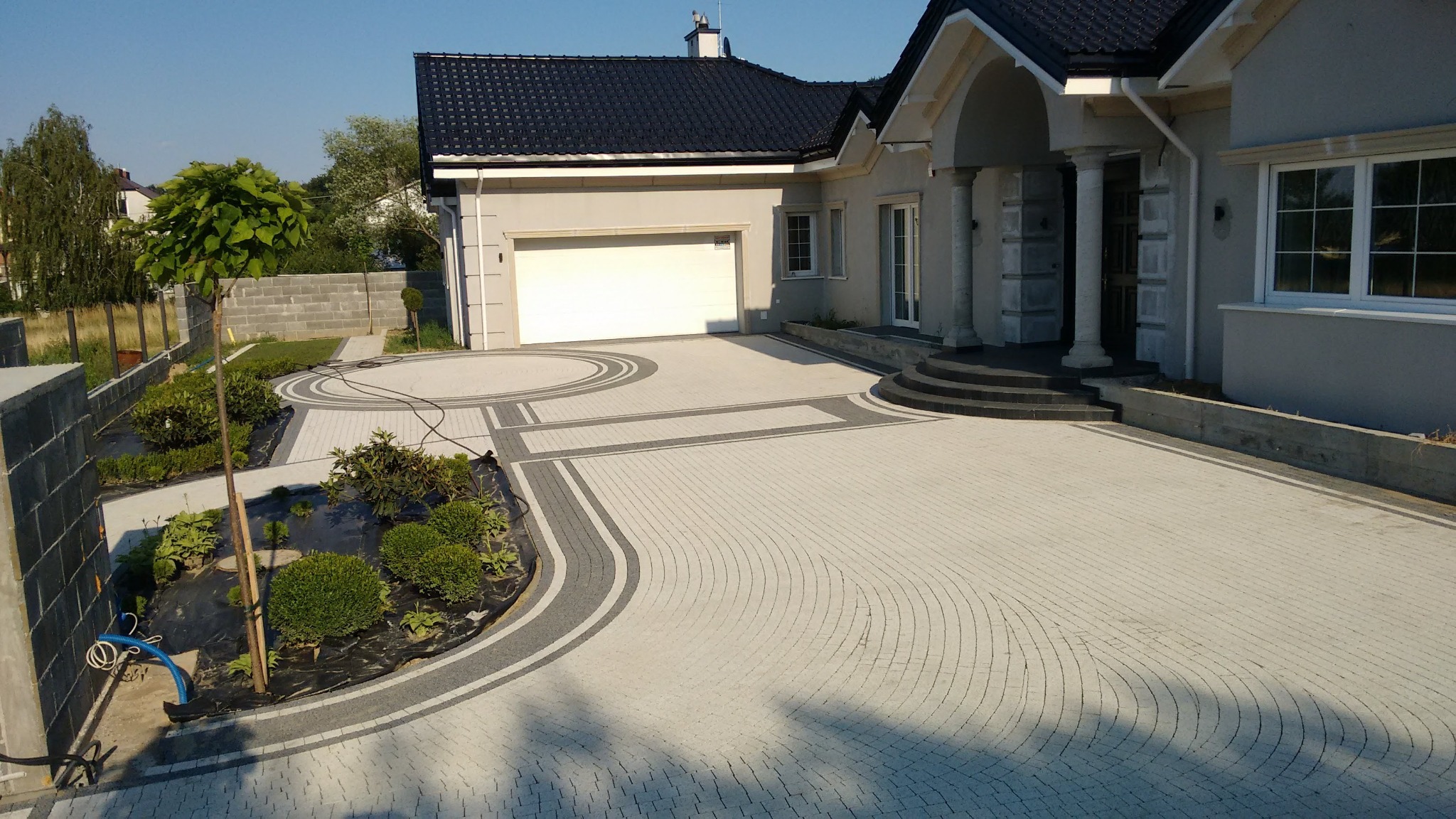 Jasnoszara kostka brukowa ułożona w łukowy wzór na podjeździe do domu z garażem, z ciemnoszarymi obramowaniami i rabatą z młodymi roślinami po lewej stronie.
