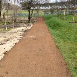 Utwardzona, brązowa droga gruntowa z widoczną siatką stabilizującą, prowadząca przez park w kierunku zabudowań mieszkalnych, z nieutwardzonym terenem po lewej stronie i trawnikiem po prawej.