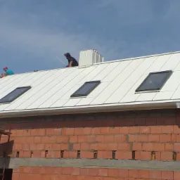 Montaż białego dachu z blachy na budynku z czerwonej cegły, widoczne okna dachowe i dwaj dekarze na dachu, jeden w czapce, drugi w kasku.