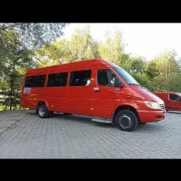 Czerwony bus Mercedes-Benz Sprinter z wysuwanym stopniem, zaparkowany na kostce brukowej, z zielonymi drzewami w tle.