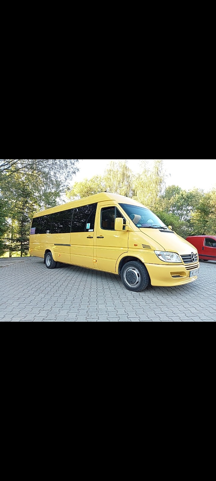 Żółty bus marki Mercedes-Benz Sprinter z przyciemnianymi szybami, zaparkowany na kostce brukowej, widok z boku.