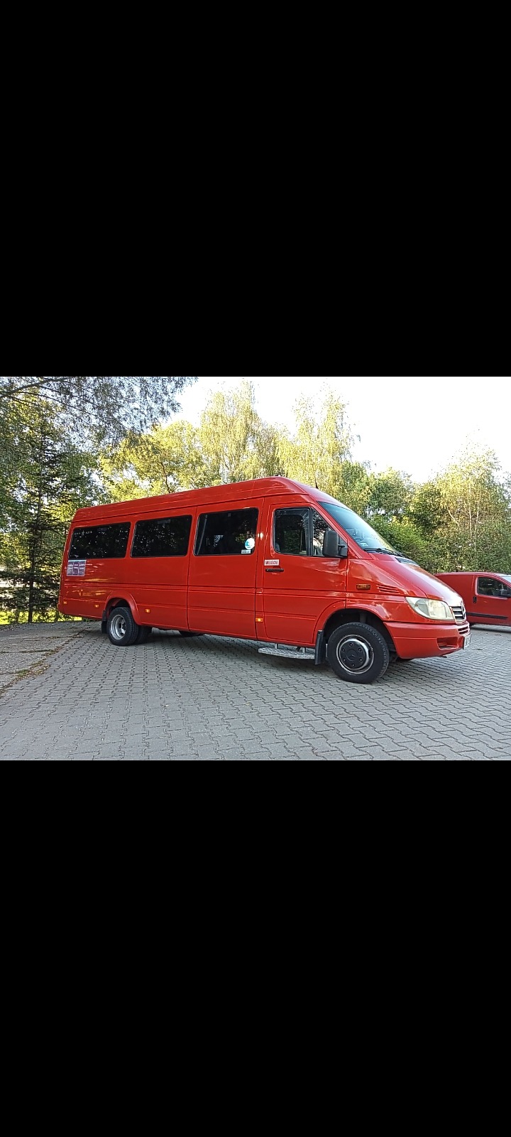 Czerwony bus Mercedes-Benz Sprinter z wysuwanym stopniem, zaparkowany na kostce brukowej, z zielonymi drzewami w tle.