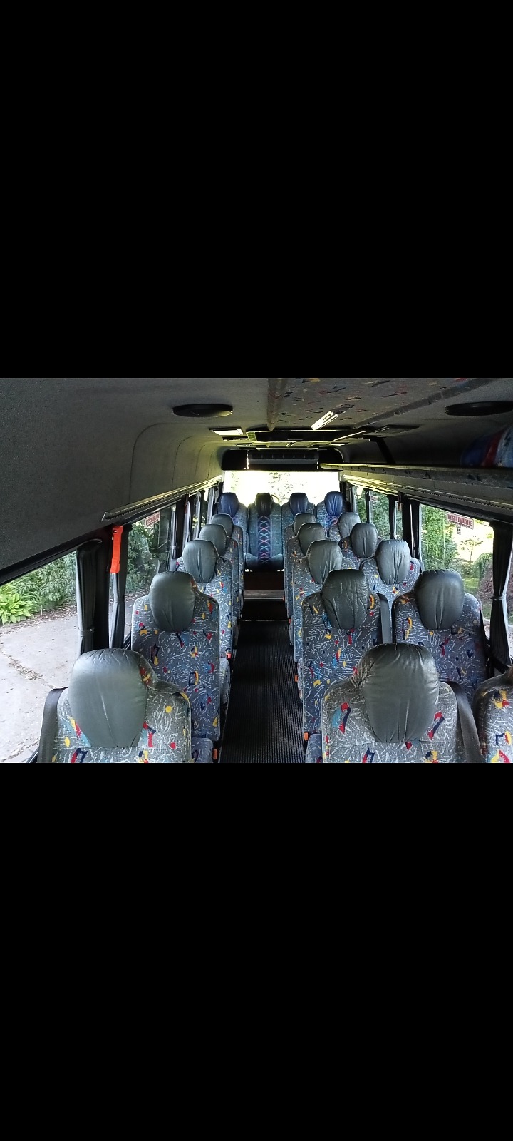 Wnętrze autobusu z rzędami siedzeń z szarą tapicerką w kolorowe wzory, widok z przodu pojazdu na puste fotele.
