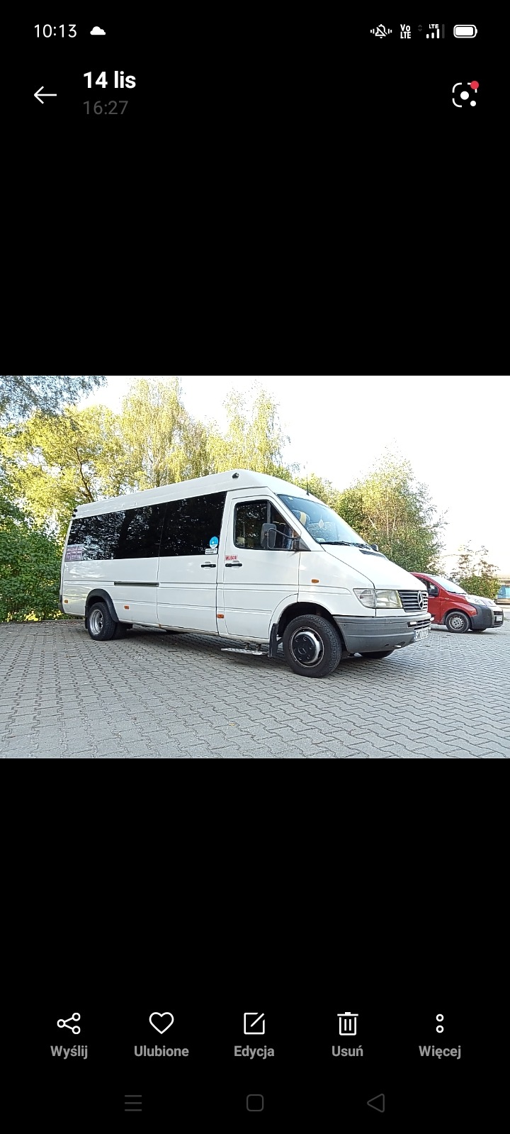 Biały bus pasażerski marki Mercedes-Benz Sprinter na parkingu z kostki brukowej, widoczny z boku, z drzewami w tle i czerwonym samochodem w oddali.