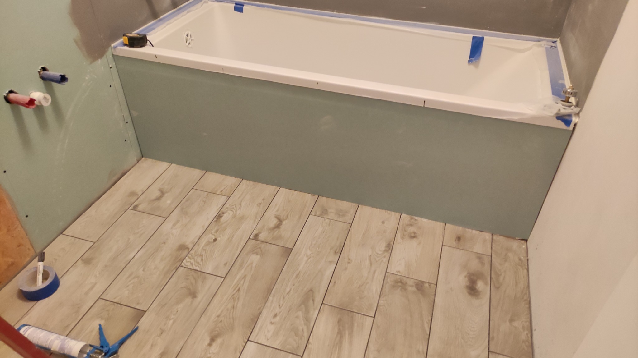 Wanna obudowana płytą gipsową, przygotowana do wykończenia, z położonymi płytkami imitującymi drewno na podłodze. Widoczne narzędzia i materiały budowlane.
