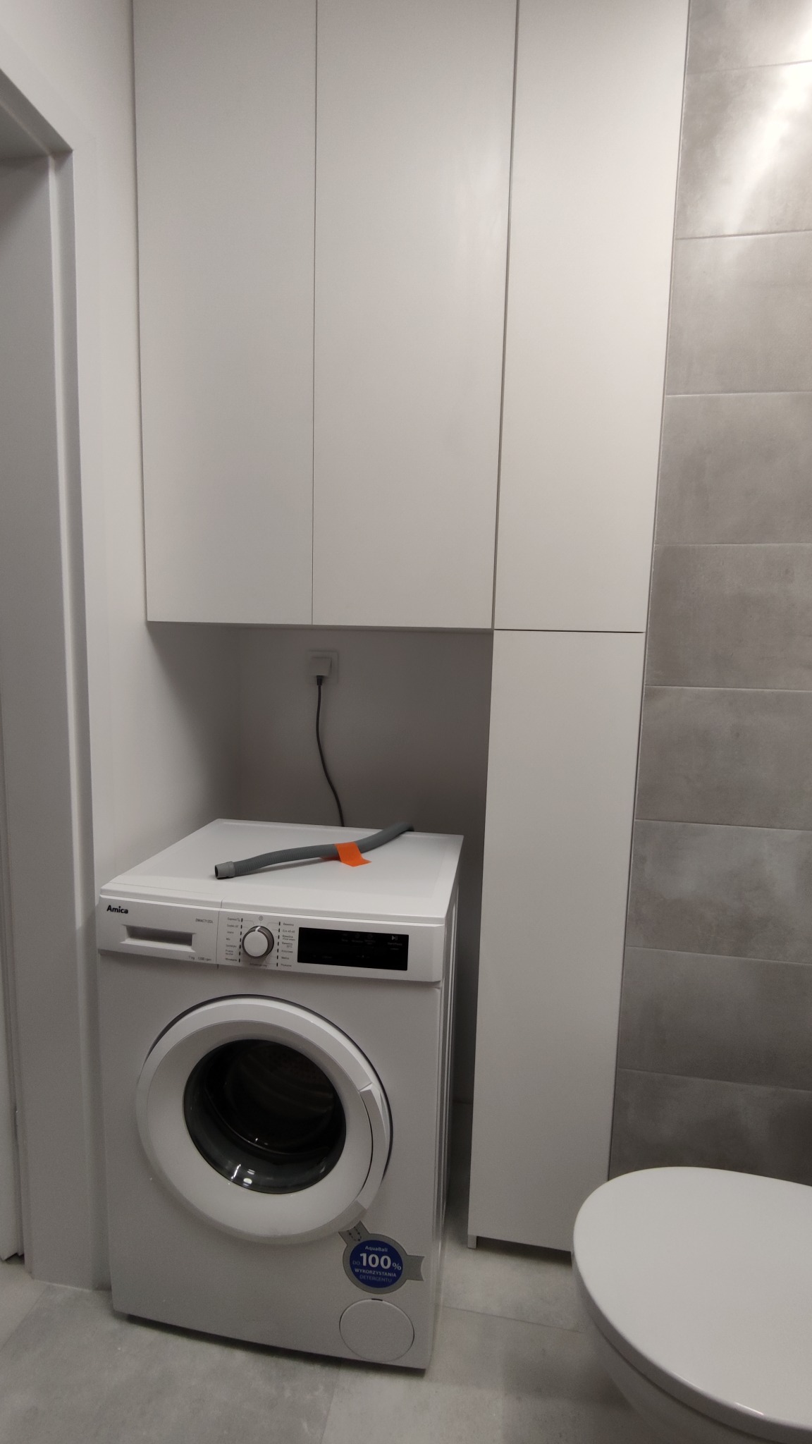 Biała pralka Amica z szarym wężem i pomarańczową etykietą na górze, umieszczona pod wiszącymi białymi szafkami w łazience z szarymi płytkami.