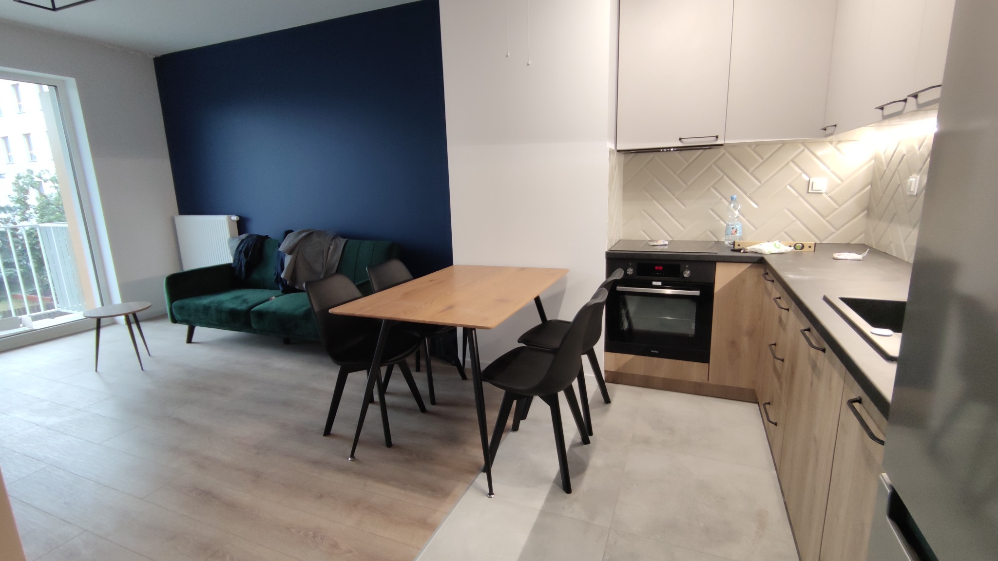 Jasne wnętrze mieszkania z aneksem kuchennym, drewnianym stołem i zieloną sofą na tle granatowej ściany, widok na ułożoną w jodełkę glazurę nad blatem.