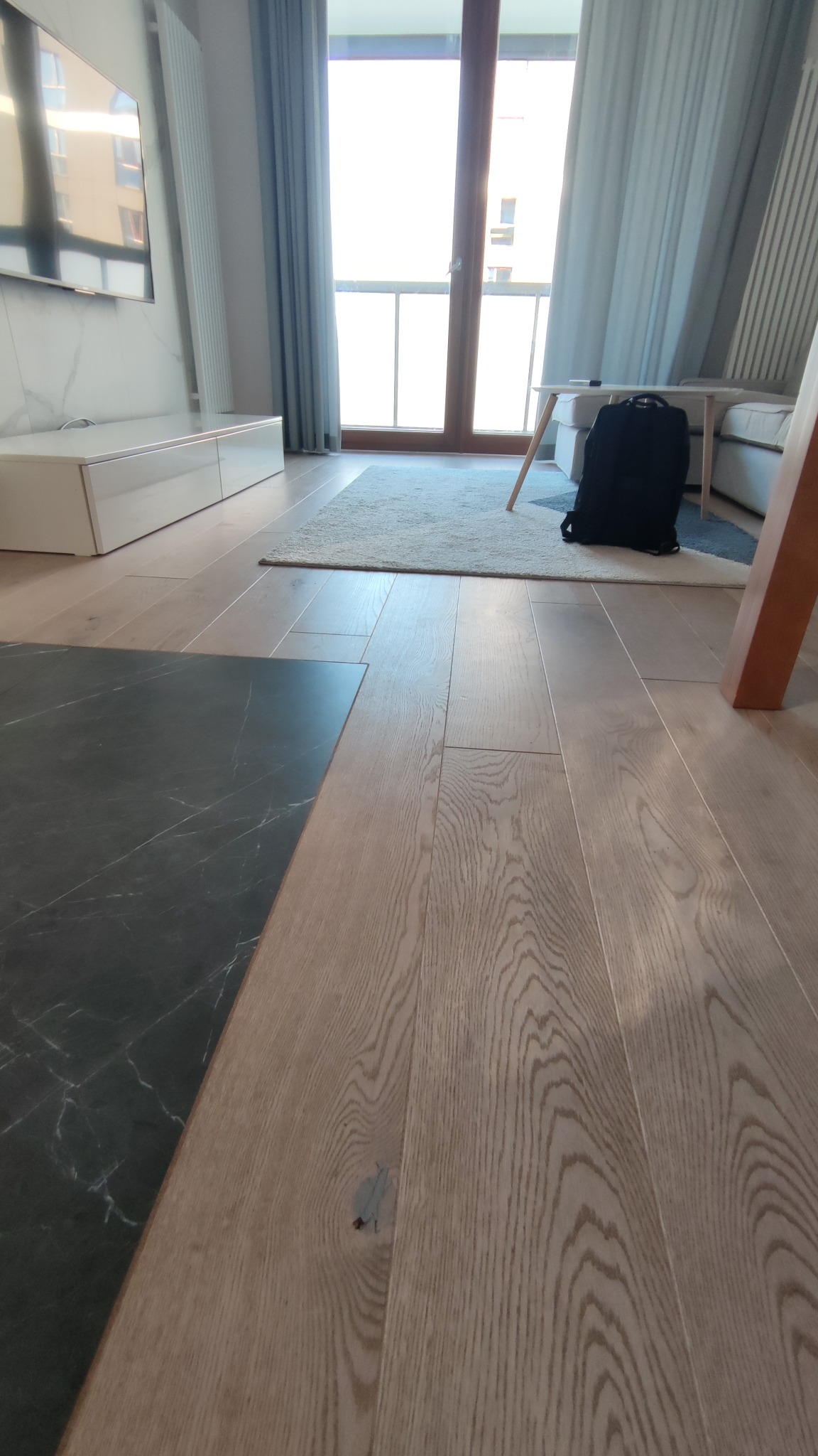 Ułożona podłoga z jasnych desek w mieszkaniu w Warszawie, z fragmentem ciemnej marmurowej płytki i widocznym fragmentem salonu z dywanem i plecakiem.