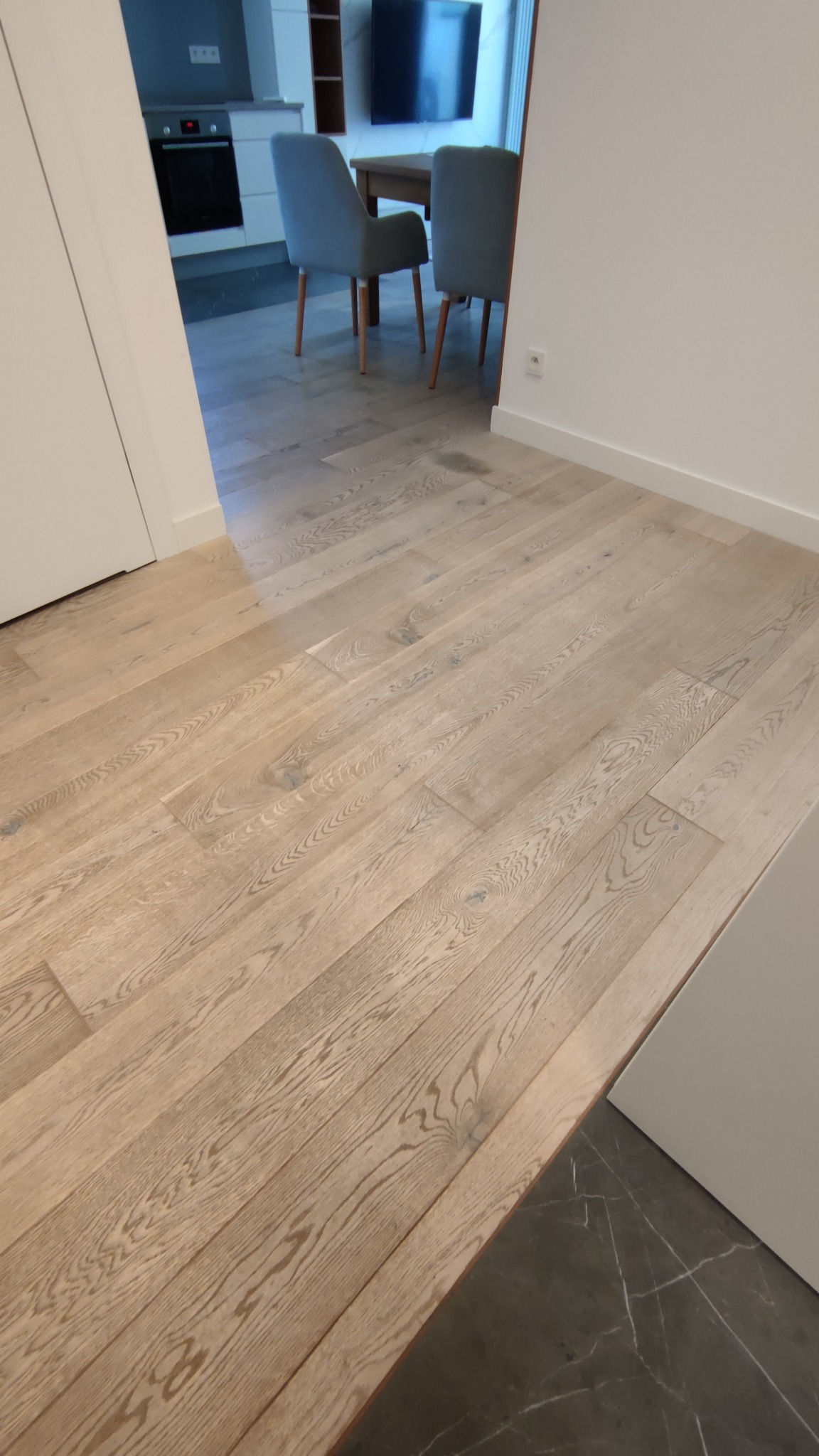 Jasna, drewniana podłoga ułożona wzdłużnie, widoczna z perspektywy podłogi, z fragmentem czarnej marmurowej podłogi w tle oraz widokiem na jadalnię z szarymi krzesłami i kuchnią w oddali.