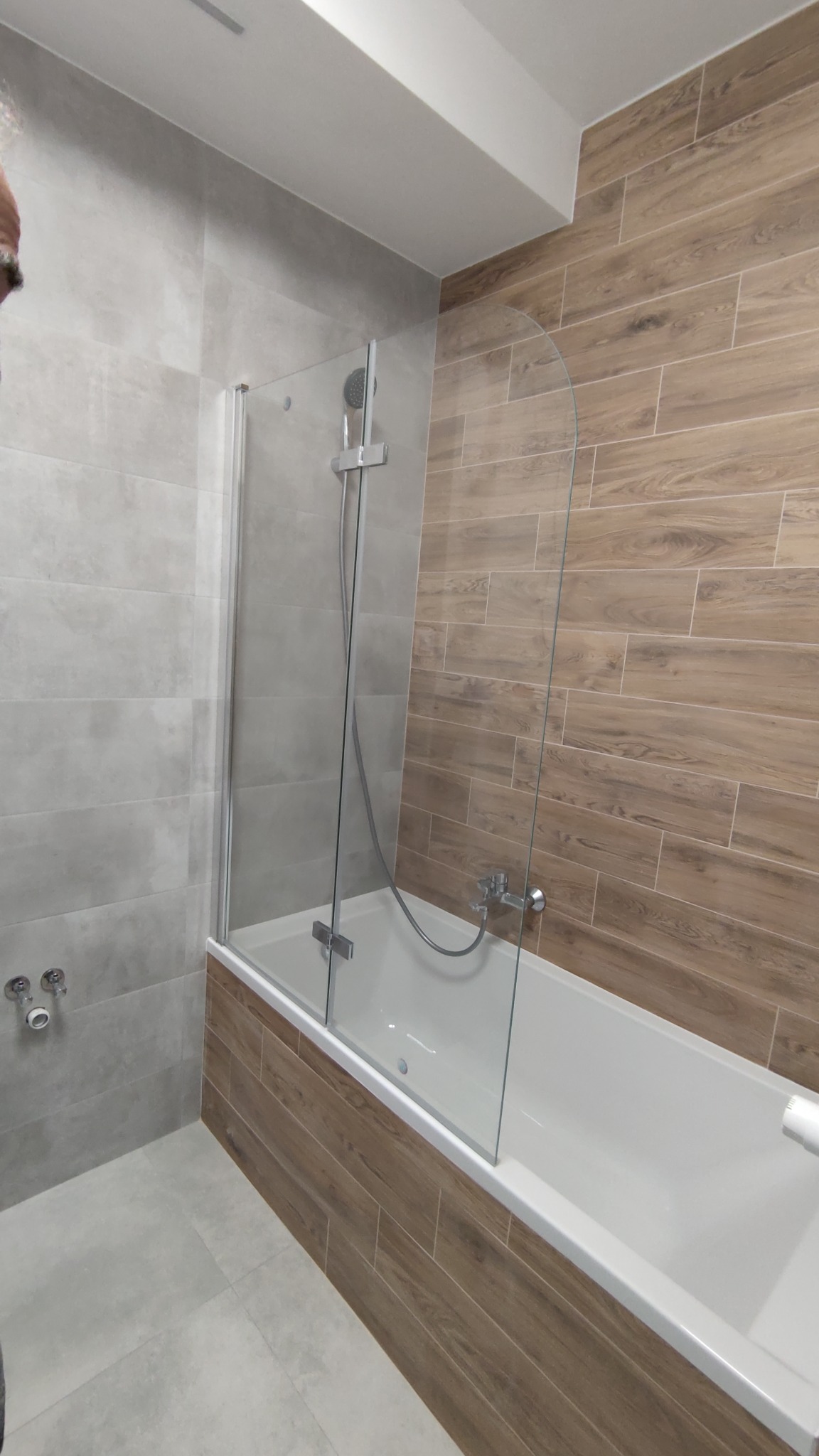 Wykończona łazienka z wanną obudowaną drewnopodobnymi płytkami, szklanym parawanem nawannowym i szarymi płytkami na ścianie, widoczne baterie i słuchawka prysznicowa.
