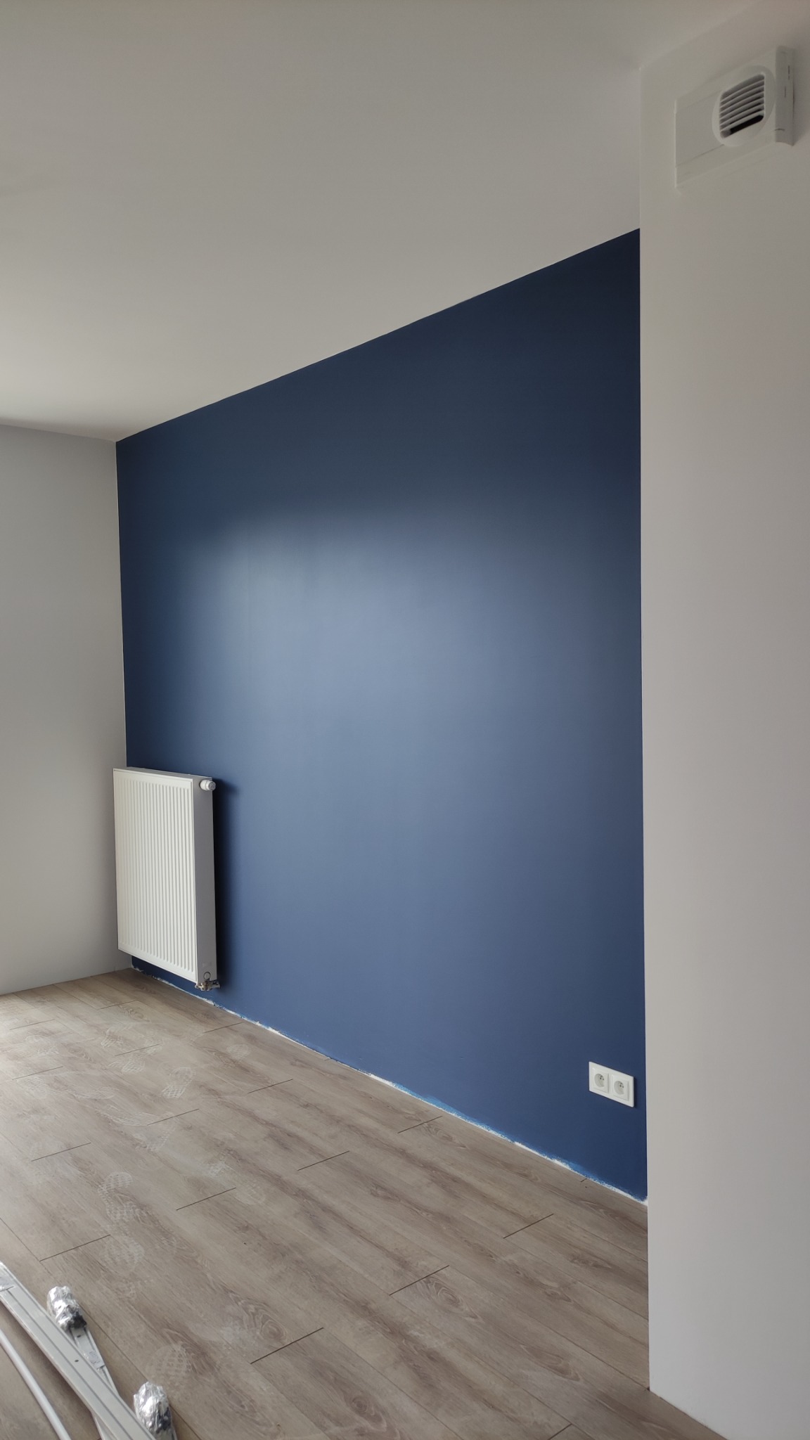 Świeżo pomalowana ściana w odcieniu głębokiego granatu w pokoju z panelami podłogowymi, widoczny biały grzejnik i gniazdko elektryczne przy listwie przypodłogowej.