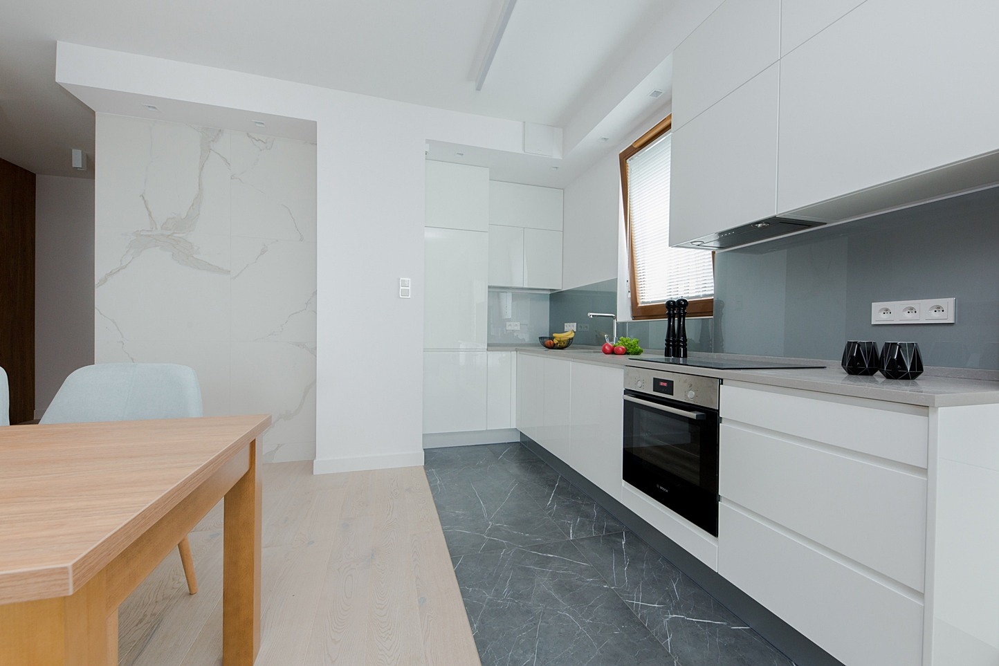 Nowoczesna, jasna kuchnia z białymi, lakierowanymi frontami szafek, szarym blatem i marmurową podłogą. Widoczny drewniany stół i krzesła w części jadalnej. Na blacie kuchennym przyprawy i owoce.