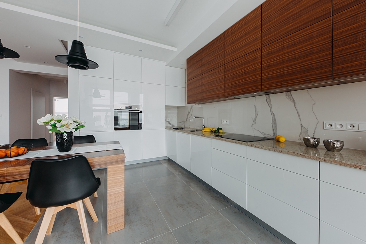 Nowoczesna kuchnia z białymi, błyszczącymi szafkami, blatem z jasnego kamienia i drewnianymi akcentami, oświetlona przez designerskie, czarne lampy wiszące.