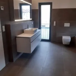 Nowoczesna łazienka z dużymi, szarymi płytkami podłogowymi i ściennymi, wisząca szafka pod umywalką z białą, prostokątną umywalką, podświetlane lustro i drzwi balkonowe z czarną ramą.